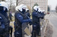 Fotografia kolorowa. Na zdjęciu widać umundurowanych policjantów zabezpieczających mecz piłkarski.