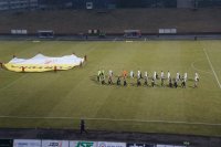 Fotografia kolorowa. Na zdjęciu widać stadion piłkarski, rozpoczęcie meczu oraz piłkarzy obu drużyn.