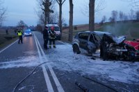 Fotografia kolorowa. Na zdjęciu widać samochód, który w wyniku wypadku uległ spaleniu oraz służby ratunkowe.