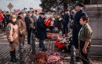 Zdjęcie kolorowe, przedstawiające składanie kwiatów przed pomnikiem.