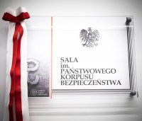 Zdjęcie przedstawiające tabliczkę z napisem oraz wstążkę w kolorze biało-czerwonym.