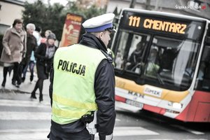 Zdjęcie przedstawiające policjanta ruchu drogowego oraz widoczny w tle autobus oraz osoby korzystające z przejścia dla pieszych.