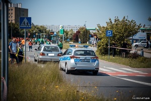 Zdjęcie przedstawiające radiowóz ruchu drogowego oraz uczestniczki biegu i osoby zabezpieczające trasę.