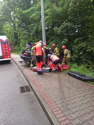 Zdjęcie przedstawiające miejsce zdarzenia drogowego, na zdjęciu widoczni strażacy, ratownicy medyczni oraz uszkodzony samochód oraz wóz strażacki.