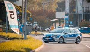 Zdjęcie przedstawiające radiowóz w trakcie zabezpieczenia oraz osoby biegające, przygotowujące się do udziału w biegu.