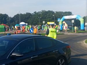 Zdjęcie przedstawiające policjanta, radiowóz w trakcie zabezpieczenia oraz osoby przygotowujące się do biegu.