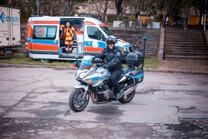 Zdjęcie przedstawiające policjanta na motocyklu oraz w oddali widoczna karetka oraz ratownik medyczny.