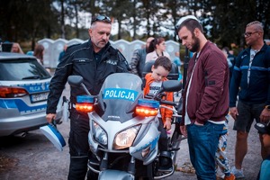 Zdjęcie przedstawiające policjanta ruchu drogowego, obok na motocyklu siedzi mały chłopiec oraz inni uczestnicy pikniku.