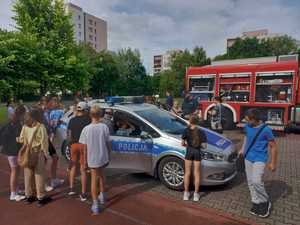 Zdjęcie przedstawiające policjanta, dzieci oraz radiowóz policyjny i wóz strażacki.