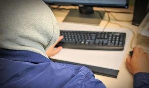Zdjęcie mężczyzny z kapturem na głowie, siedzącego przy komputerze.